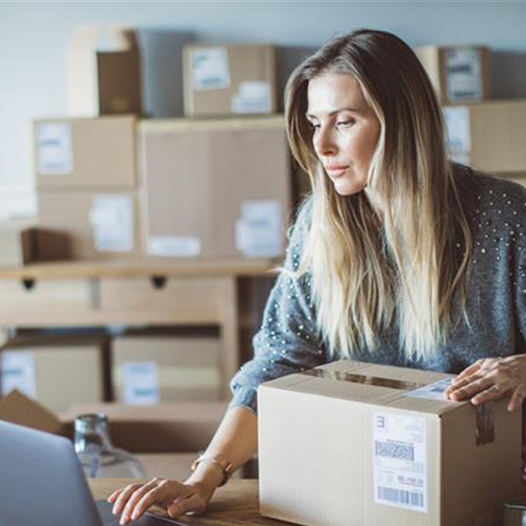 Woman with one hand on a package on a desk and the other using a laptop with lots of packages behind her