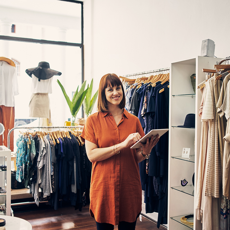 A woman in an orange shirt dress stands in a clothing store, using a tablet, surrounded by racks of stylish clothing and accessories.