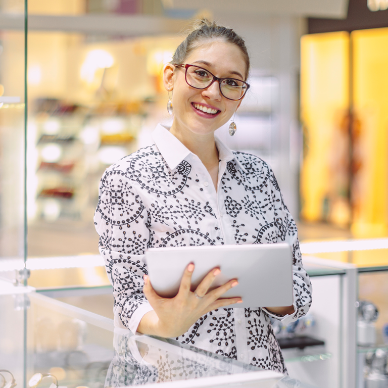 A retail assistant standing behind a low display case filled with watches, holding a tablet, using AI in retail and engaging with customers in a modern shopping environment.