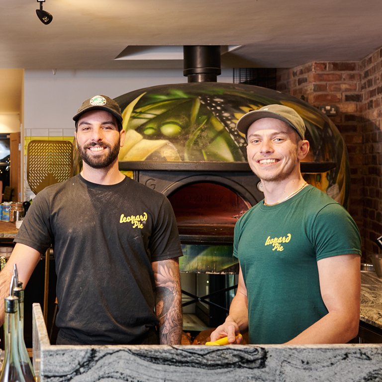 Staff members preparing pizzas in a vibrant kitchen environment, with stacks of pizza boxes and a traditional pizza oven in the background.