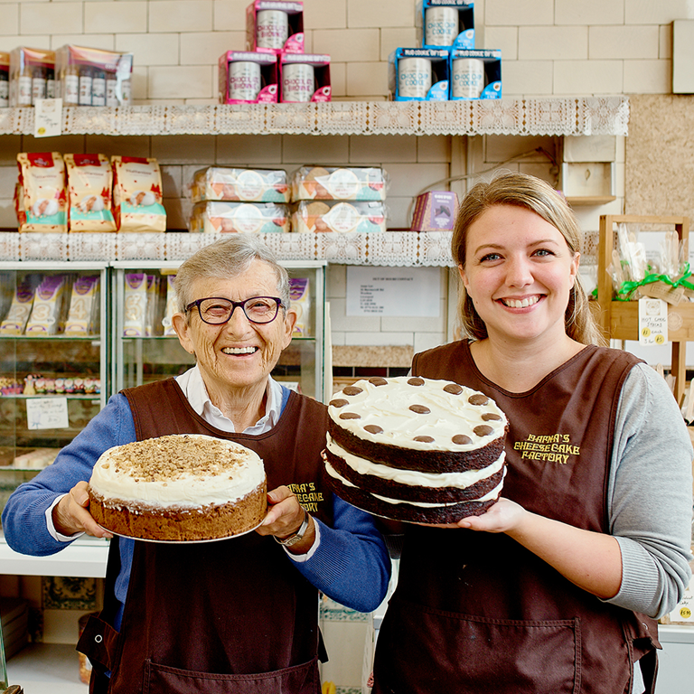 Two women in a bakery holding large cakes, surrounded by a display of sweets and baked goods, representing the importance of succession planning in family businesses.