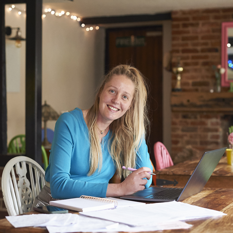 A woman in a blue sweater is working on a laptop at a wooden table in a cozy room with colorful chairs and a brick fireplace, illustrating the concept of making tax digital in a comfortable home office setting