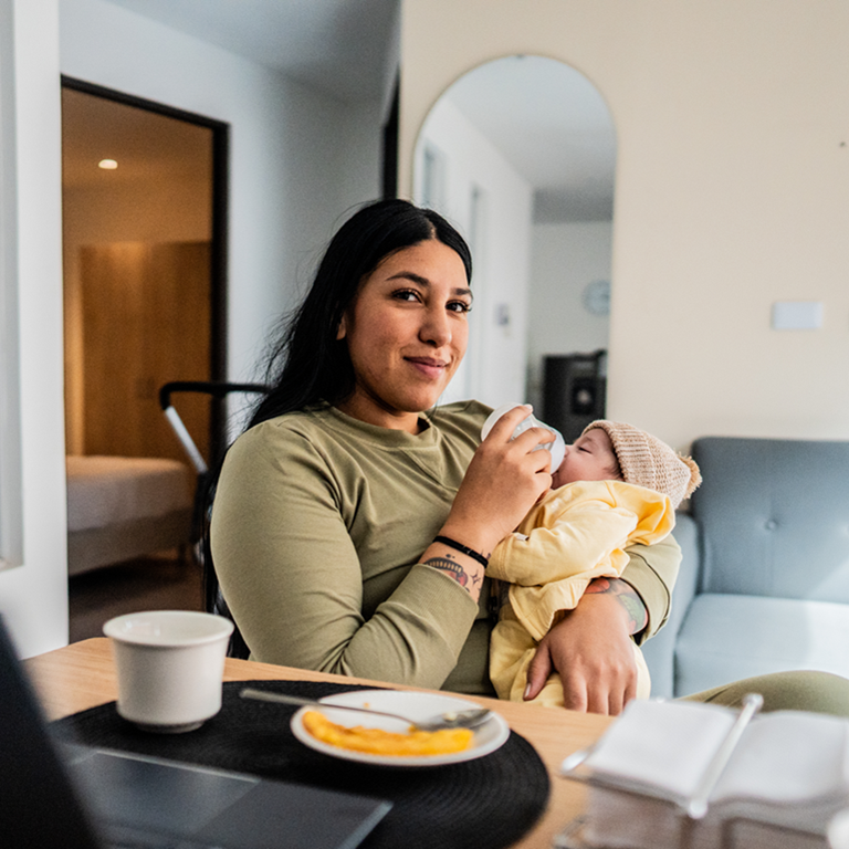 A mother holding and feeding her baby with a bottle while sitting at a table with a laptop, cup, and plate in a cozy home setting