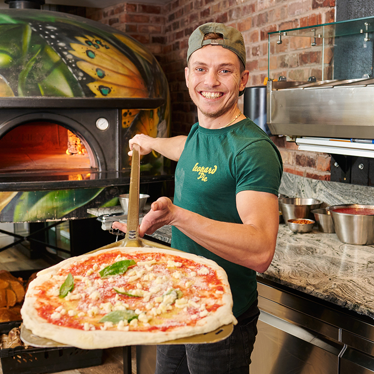 Leopard Pie pizza restaurant team member holding uncooked pizza on a pizza peel in front of pizza oven 