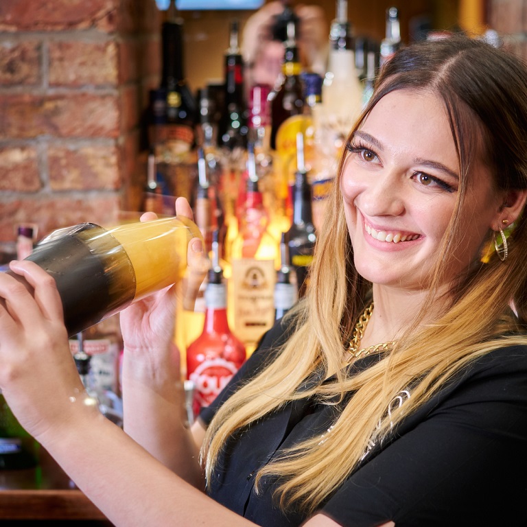 Smiling female bartender shaking a cocktail with lots of drinks bottles behind her